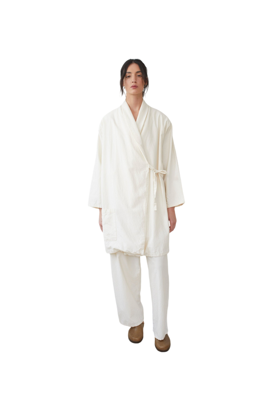 Dressed in Wol Hide's "Twill Tie Trench: Natural," the individual stands against a plain black background, showcasing a loose-fitting kimono-style outfit crafted from organic cotton. The ensemble, paired with light brown shoes, features a robe and wide-legged pants.