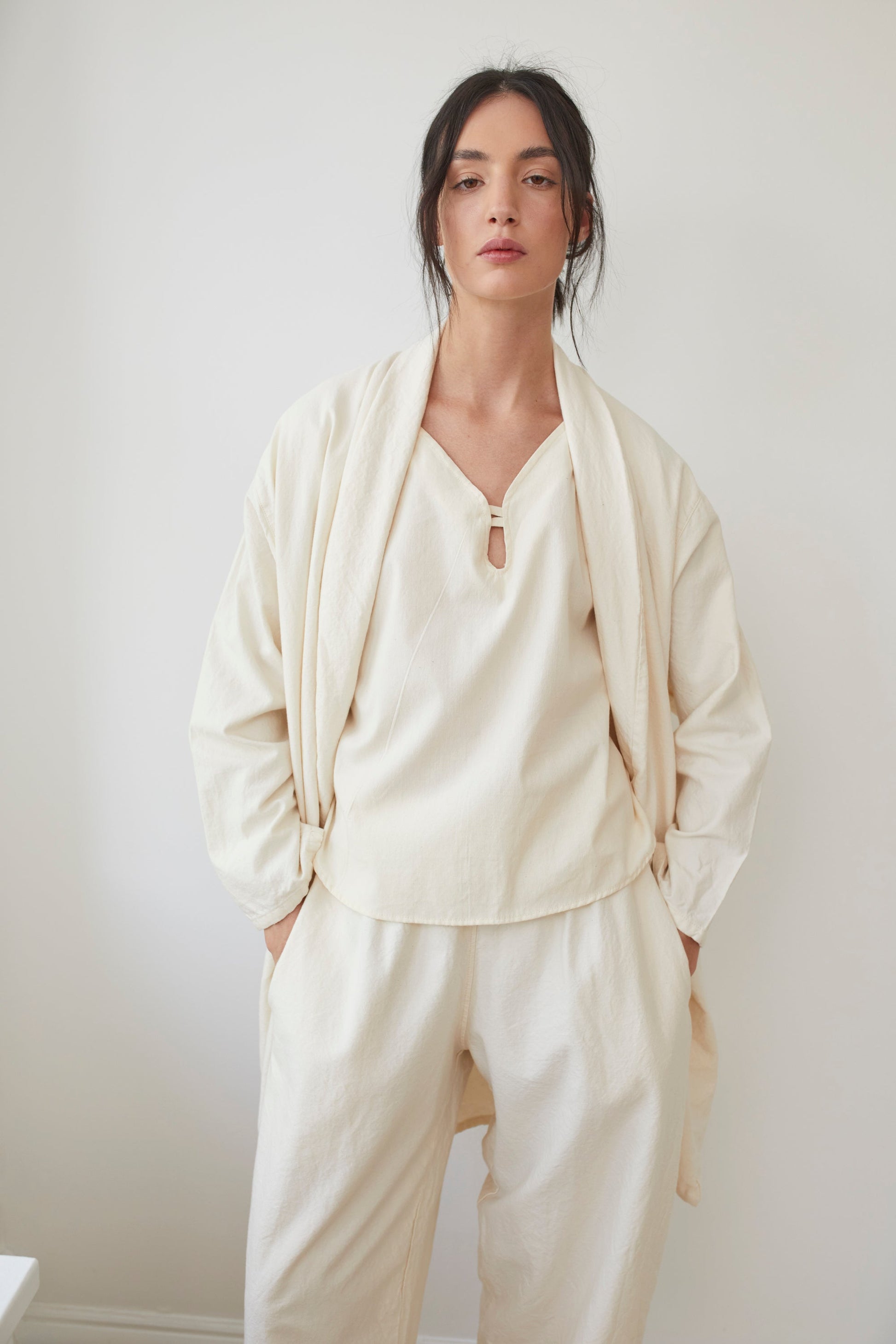 A person with dark hair stands against a light-colored wall, wearing the loose-fitting Twill Tie Trench in Natural by Wol Hide, made in the USA. This ensemble features an oversized jacket and relaxed pants crafted from organic cotton, creating a simple and elegant look.