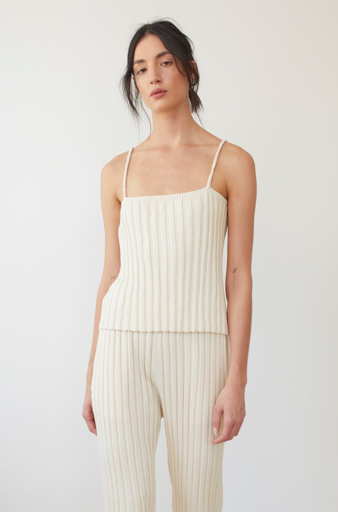 A person poses against a simple background dressed in the Rib Cami : Ecru by Wol Hide—an exquisite cream-colored, ribbed, sleeveless top featuring braided straps. They also wear matching ribbed pants, with their dark hair styled in a loose updo, accentuating the elegance of the organic cotton camisole.