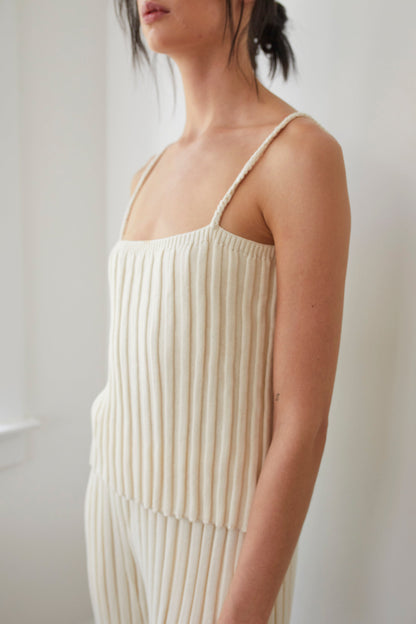 Standing near a window, a person embodies the effortless elegance of Wol Hide in their Rib Cami : Ecru, a sleeveless, ribbed knit top crafted from regenerative Peruvian cotton. The light beige garment features a texture akin to vertical stripes, with their hair tied back as they face sideways.