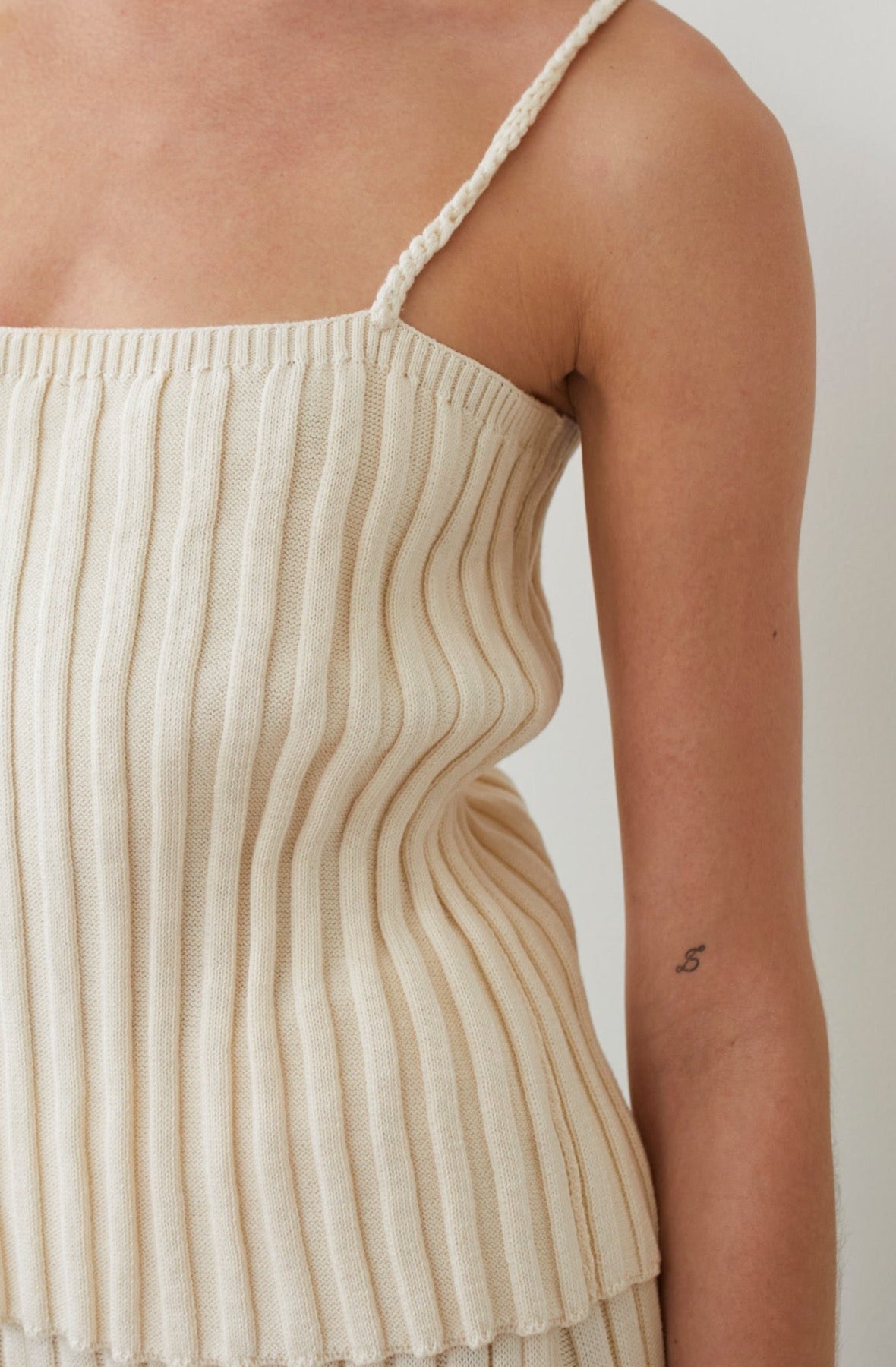 Close-up of a person wearing the Rib Cami in Ecru by Wol Hide, featuring braided straps. A small tattoo of the number "5" adorns their upper arm. Made from organic cotton, this top contrasts beautifully against the plain, neutral background.