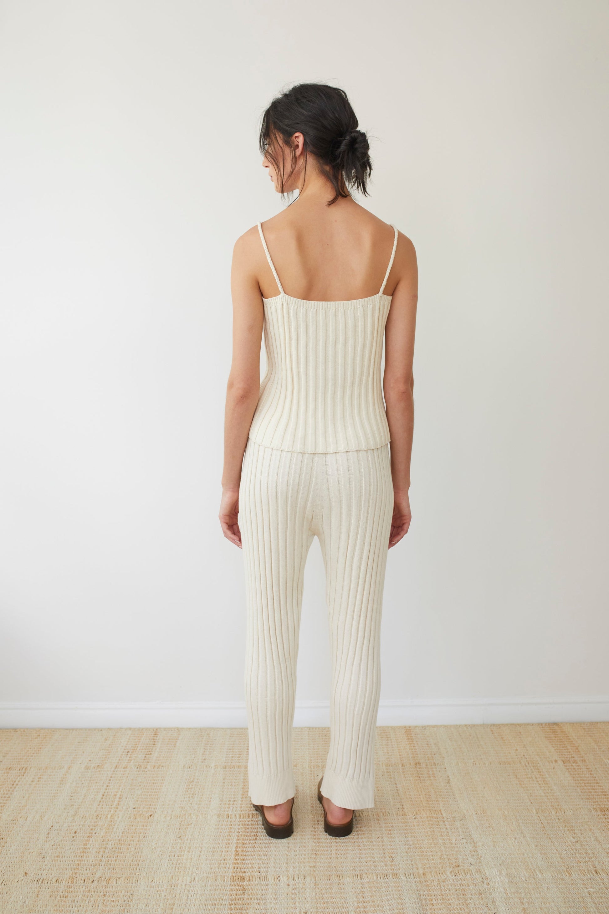 A woman with dark hair stands facing away, wearing the "Rib Cami : Ecru" by Wol Hide, a sleeveless ribbed top with braided straps. She pairs it with matching pants made from organic regenerative cotton and brown sandals. The backdrop is a plain white wall and she is on a light-colored floor.