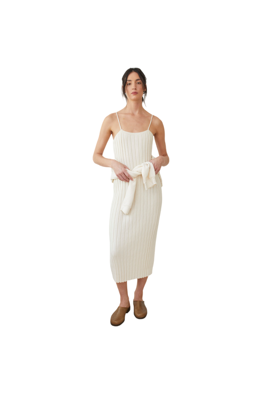 A person wearing the Rib Dress in Ecru by Wol Hide, featuring a white, sleeveless design with braided straps, is holding a cream-colored sweater tied around their waist. They have brown hair tied back and are standing against a black background, wearing brown shoes.