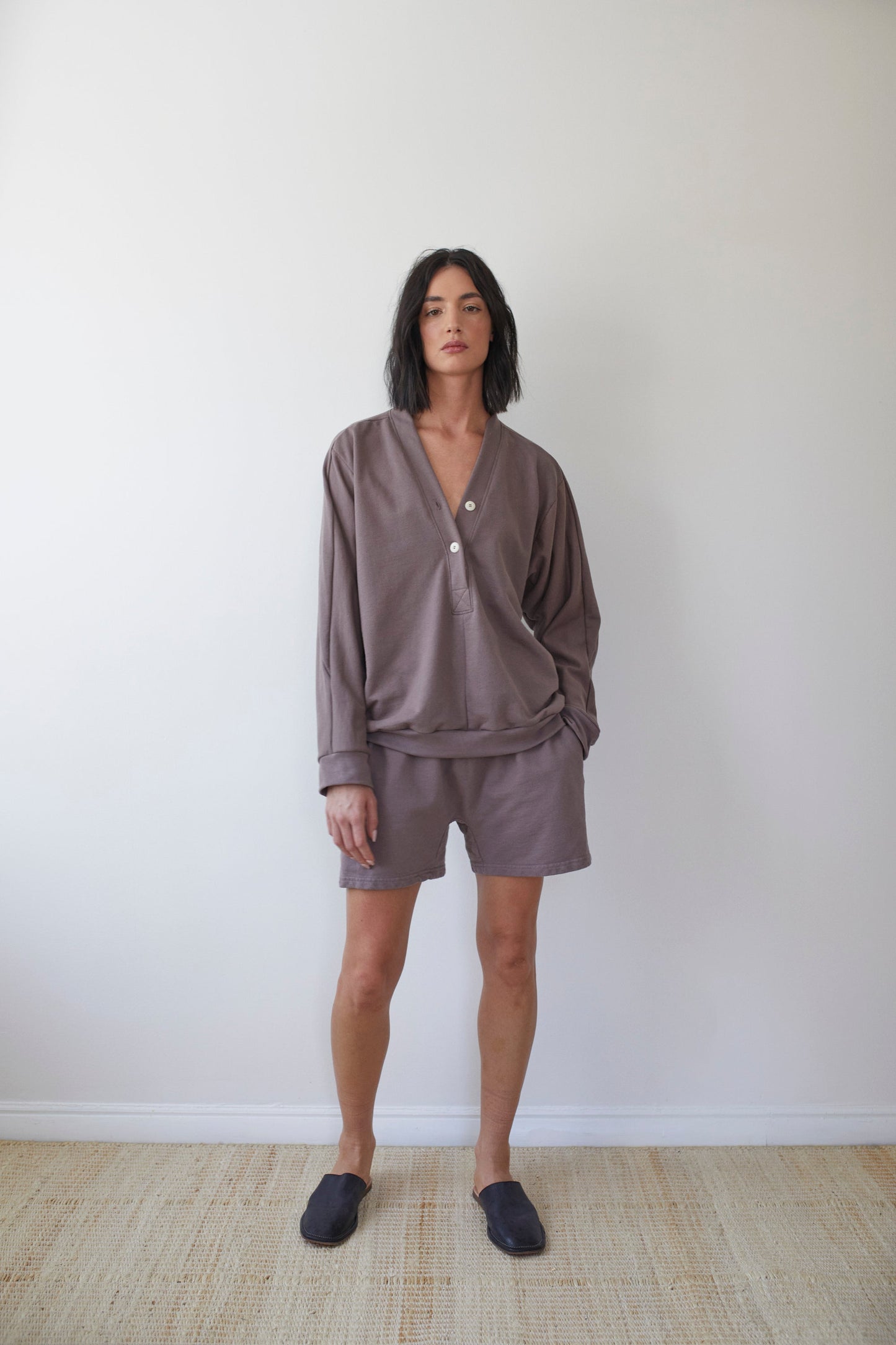 A person with shoulder-length dark hair wears a loose long-sleeve brown top paired with Wol Hide's Organic Cotton French Terry Easy Shorts in Mushroom and dark slippers. They stand against a plain white wall on a light-colored floor, gazing at the camera.