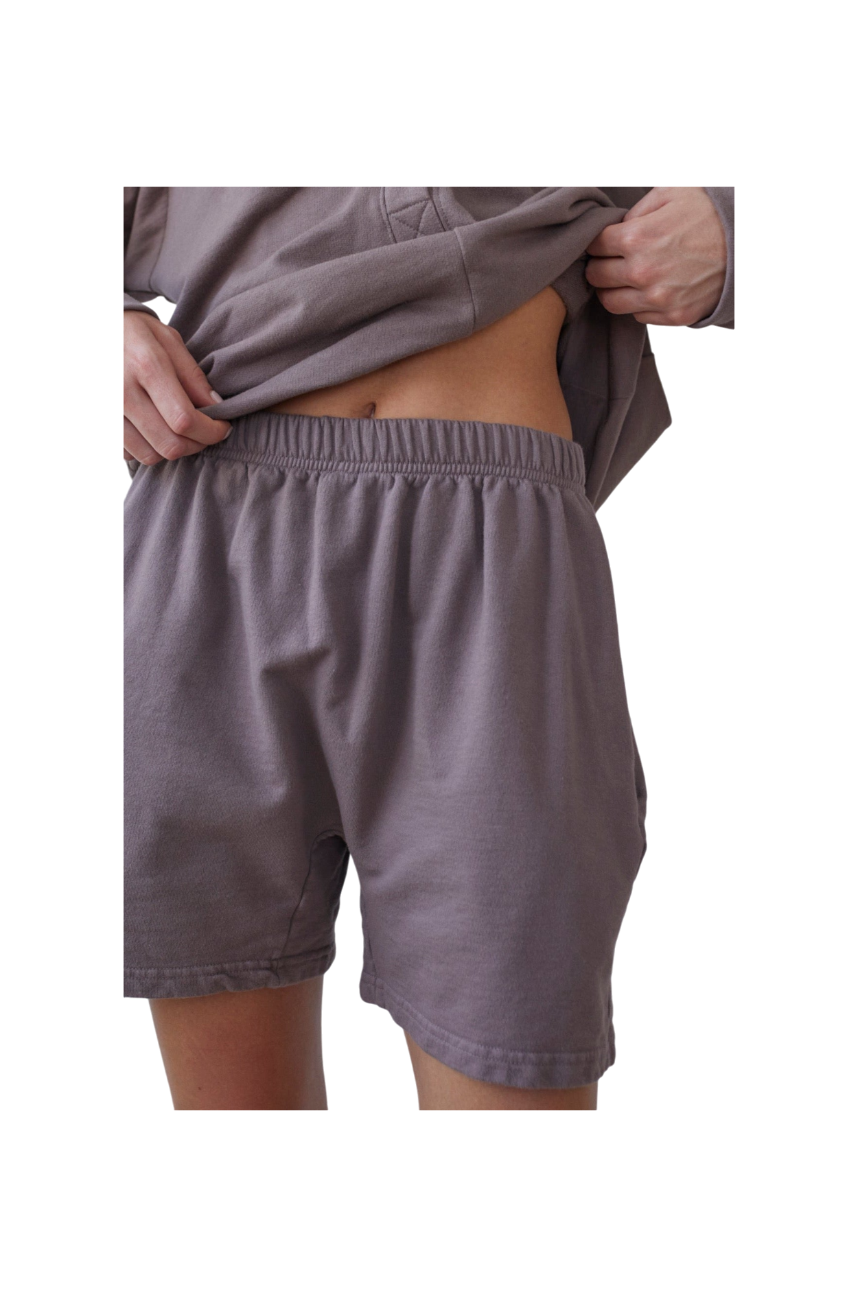 A person wearing the Wol Hide Cotton Easy Shorts in Mushroom and a matching gray sweatshirt made from Organic Cotton French Terry is lifting the bottom of the sweatshirt to reveal a small portion of their abdomen. The image focuses on the midsection and part of the legs and arms, with a black background.