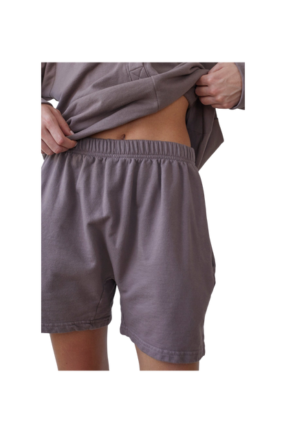 A person in a loose long-sleeve shirt and Wol Hide's Organic Cotton French Terry Easy Shorts in Mushroom gently lifts their shirt to reveal their midriff. The USA-made shorts create a striking contrast against the black background.