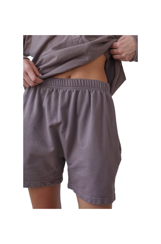 A person wearing the Wol Hide Cotton Easy Shorts in Mushroom and a matching gray sweatshirt made from Organic Cotton French Terry is lifting the bottom of the sweatshirt to reveal a small portion of their abdomen. The image focuses on the midsection and part of the legs and arms, with a black background.