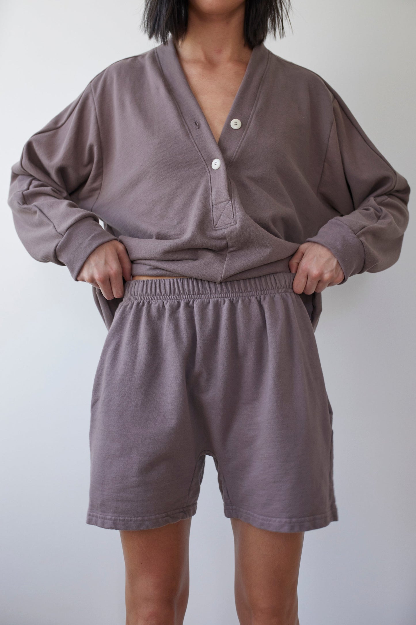 A person is wearing a loose-fitting, long-sleeve shirt featuring a V-neck and buttons, paired with Wol Hide's comfortable Cotton Easy Shorts in Mushroom. Both garments are crafted from soft Organic Cotton French Terry in a muted greyish-purple hue. The person's hands are tucked into the waistband of the shorts.