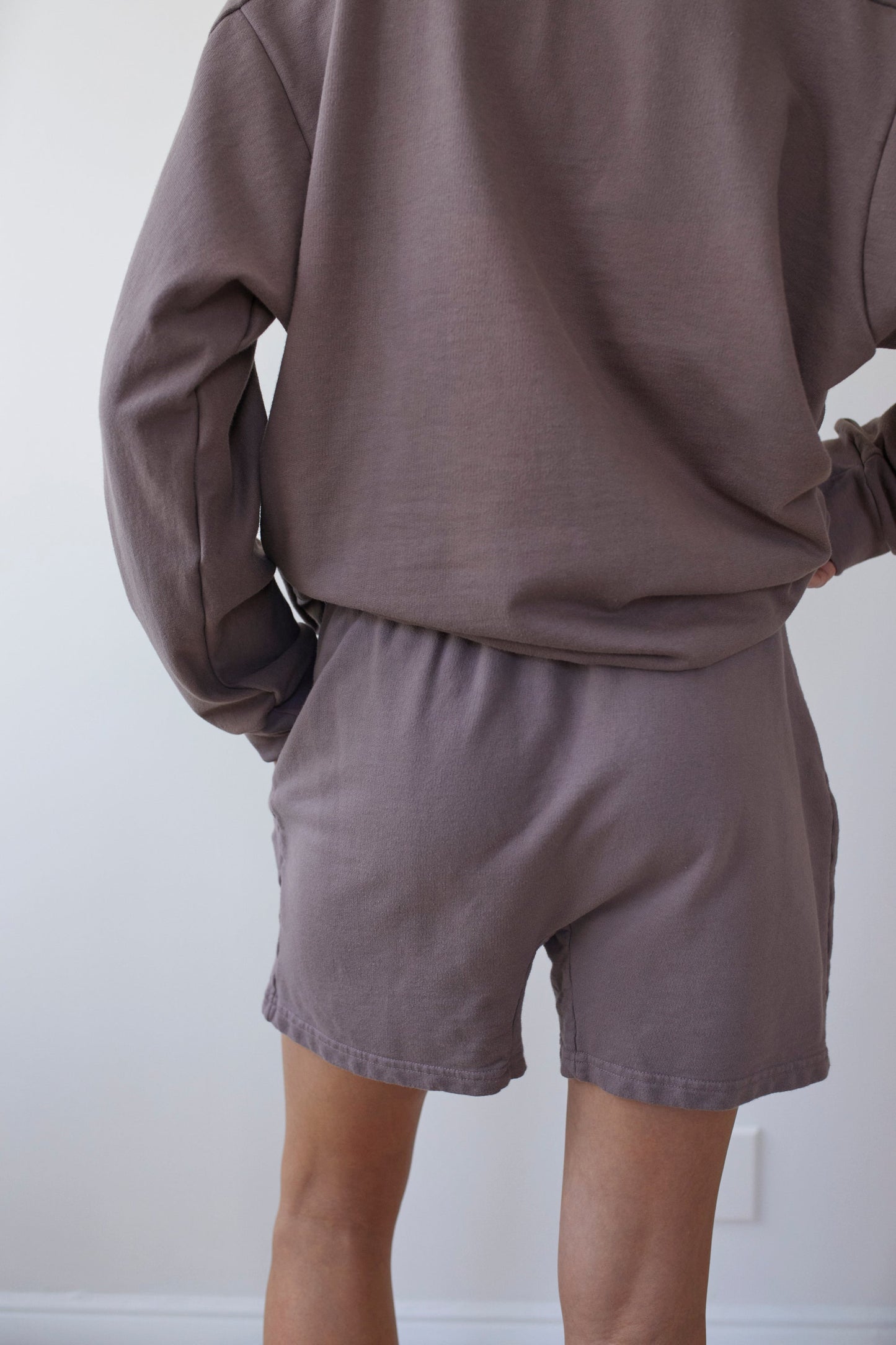 A person is shown from the back, dressed in a matching brown long-sleeve shirt and Wol Hide's Cotton Easy Shorts in Mushroom. The outfit has a relaxed, comfortable fit. The background is plain and light-colored, emphasizing the casual attire made in the USA.