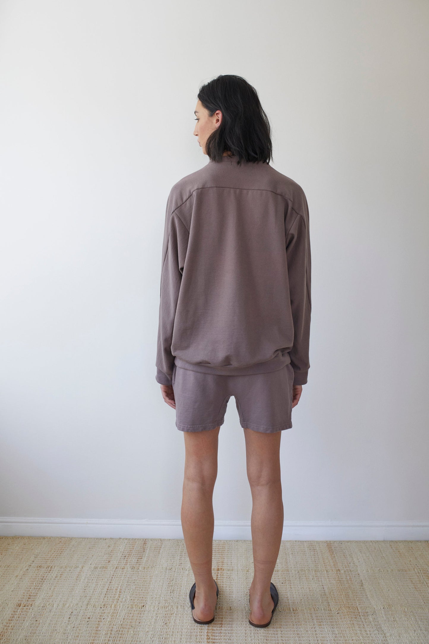 A person with shoulder-length dark hair stands indoors against a white wall, wearing Wol Hide's loose-fitting, long-sleeve brown top and Organic Cotton French Terry Easy Shorts in Mushroom. They pair the outfit with black flip-flops, standing on what appears to be a light-colored carpeted floor.