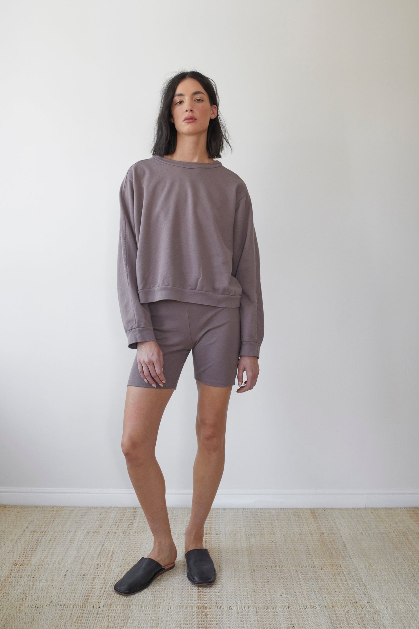 A person with long dark hair stands against a light-colored wall, wearing a loose-fit Wol Hide Cotton French Terry Easy Summer Sweatshirt in Mushroom and matching fitted grey shorts. They are also wearing dark grey slip-on shoes and standing on a light beige carpet.