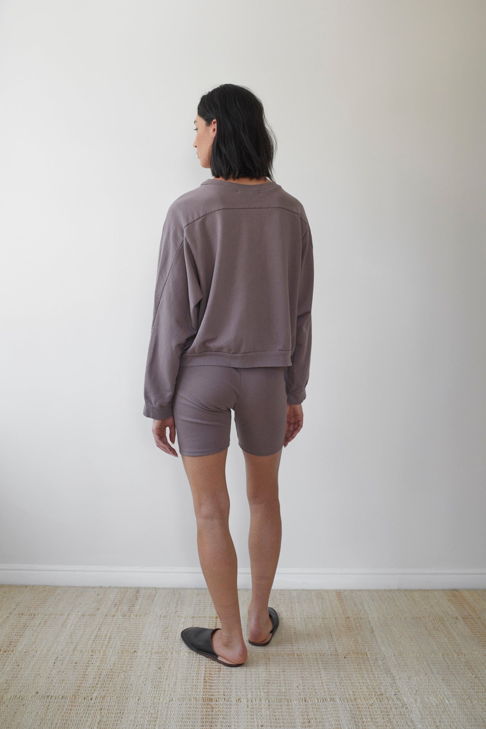 A person with shoulder-length black hair stands facing away, wearing a loose, long-sleeved top made from Organic Cotton French Terry and matching shorts, both in a muted, earthy tone. They are standing on a neutral-colored woven rug against a plain off-white wall.