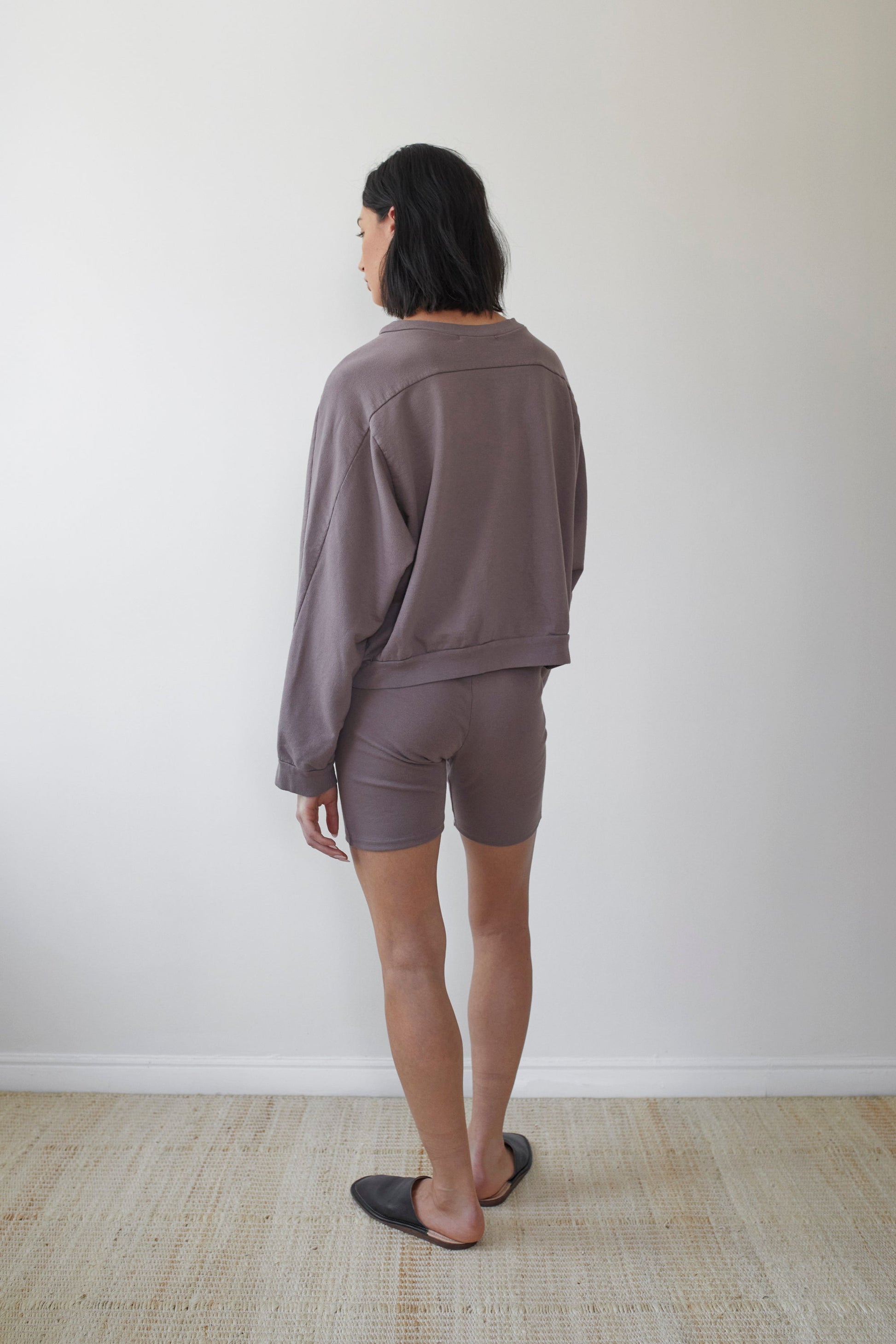 A person with shoulder-length dark hair stands facing away from the camera, wearing a loose-fitting, long-sleeved, dark gray top made of organic cotton and matching Wol Hide Cotton Bike Shorts in Mushroom. They are also wearing black slip-on shoes and are standing on a light-colored woven rug against a plain white wall.