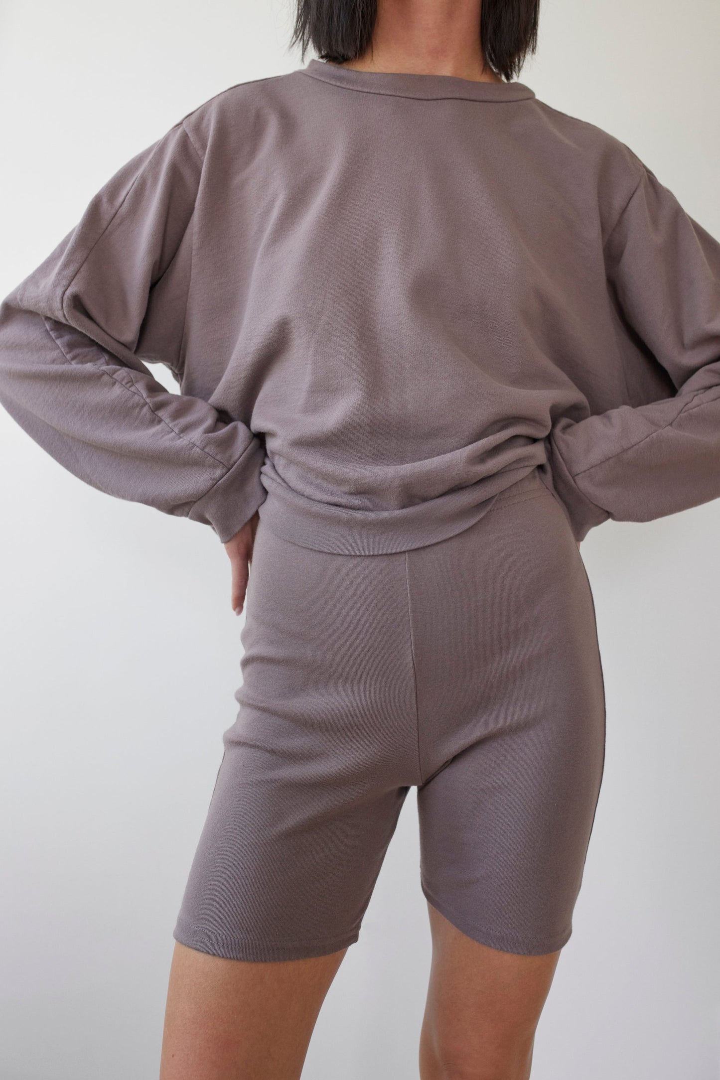 A person stands with their hands on their hips, wearing a relaxed-fit, long-sleeved shirt and fitted Cotton Bike Shorts in Mushroom by Wol Hide. The ensemble, made from organic cotton and manufactured in the USA, sits against a plain white background.