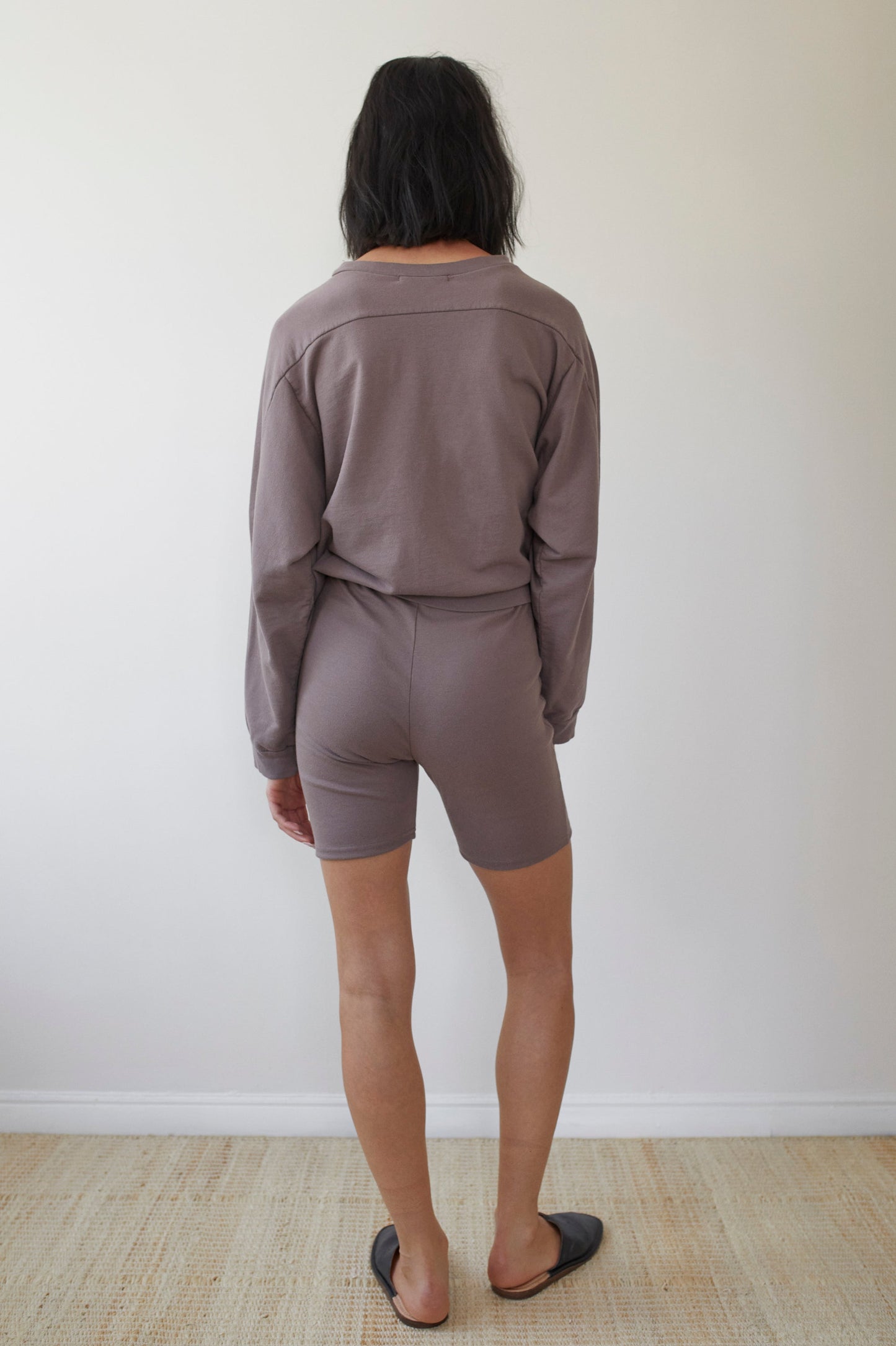 A person with shoulder-length dark hair stands facing a plain white wall, captured from the back. They are wearing matching light brown organic cotton long-sleeve top and Wol Hide's "Cotton Bike Shorts in Mushroom," paired with black slip-on shoes. The floor is light-colored wood, and the room appears to be well-lit.