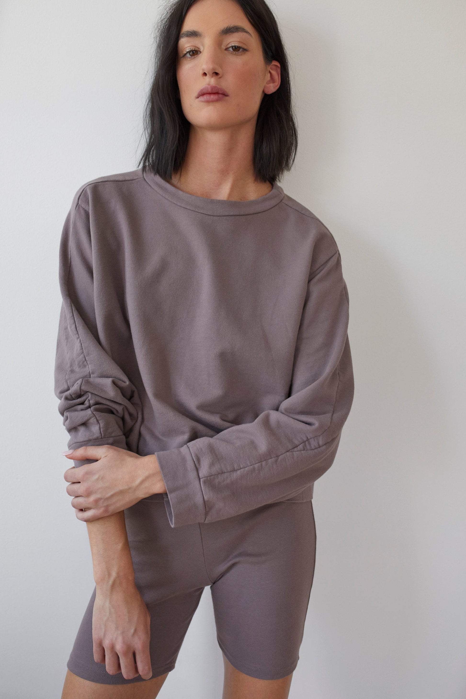 A person with medium-length black hair is leaning against a white wall. They are wearing Wol Hide's relaxed-fit, long-sleeve Cotton French Terry Easy Summer Sweatshirt in Mushroom and matching shorts, gazing directly at the camera with a neutral expression.