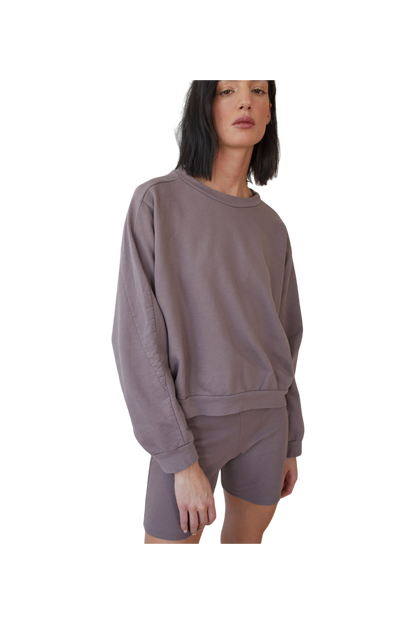 Against a simple backdrop, an individual wears Wol Hide's loose, long-sleeved Organic Cotton French Terry Easy Summer Sweatshirt in Mushroom with matching shorts. Their shoulder-length dark hair frames their face as they gaze off-camera, exuding effortless style.