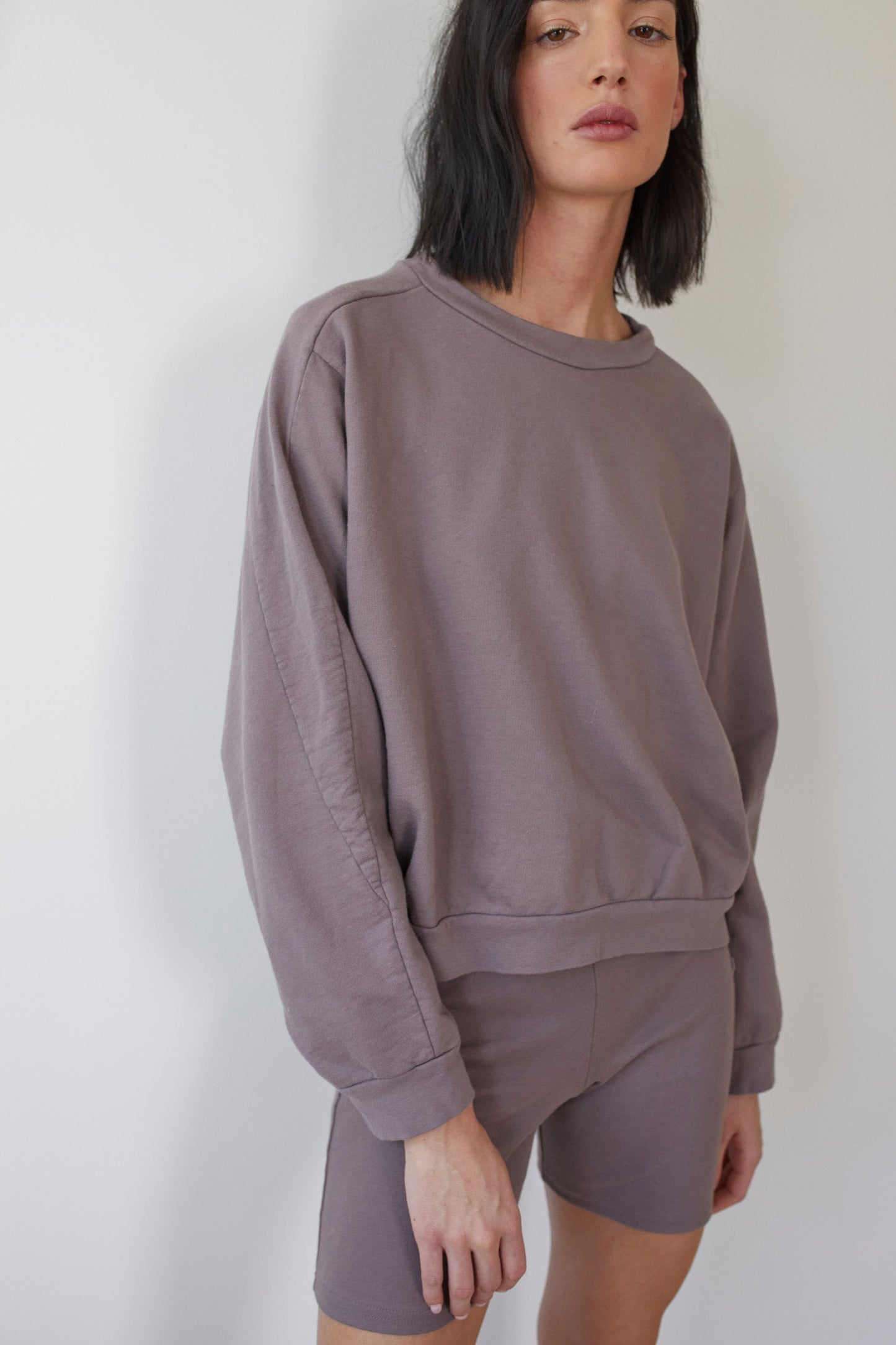 A person with medium-length black hair is wearing a casual outfit consisting of an oversized, long-sleeved Cotton French Terry Easy Summer Sweatshirt in Mushroom by Wol Hide and matching high-waisted shorts. They are standing against a plain white background.