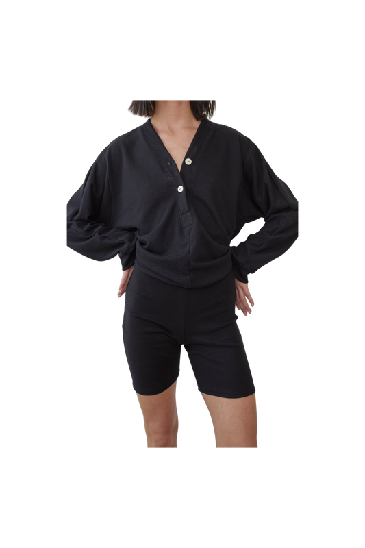 A person is standing against a plain background, wearing a black, long-sleeve button-up top with two buttons undone and high-waisted Cotton Bike Shorts in Black from Wol Hide. Their hands are placed on their hips, and their face is not visible.