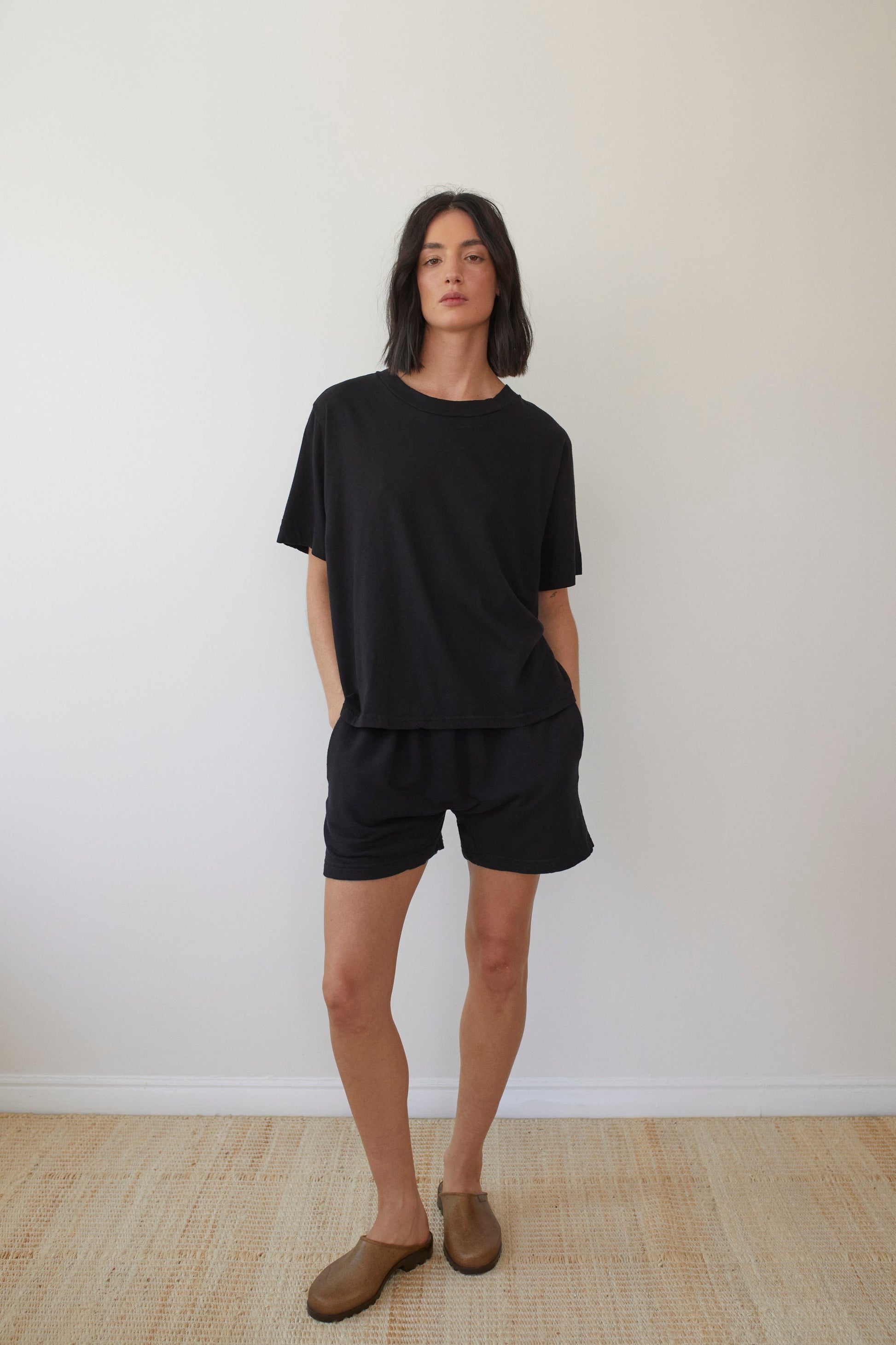 A person with short, dark hair stands against a plain white background. They are wearing the loose-fitting Wol Hide Cotton Jersey Box Tee in Black and matching black shorts, with brown slip-on shoes. The person's hands rest casually at their sides.