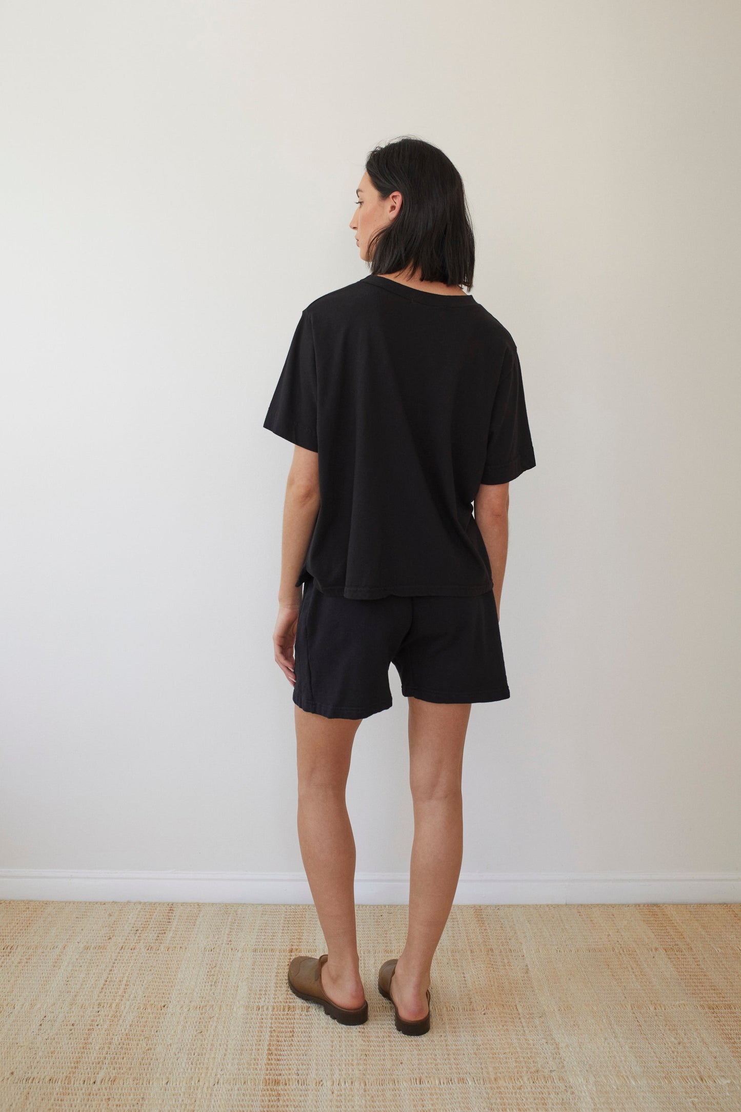 With long dark hair, a person stands in a minimalist room with light wood floors and plain white walls. They're wearing Wol Hide's Organic Cotton Jersey Box Tee in black, matching shorts, and brown slip-on shoes.