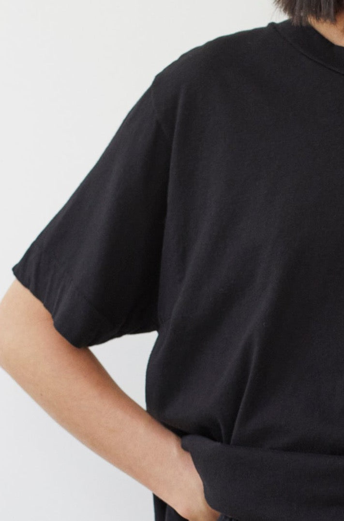 A person is shown from the shoulders down wearing the Wol Hide Cotton Jersey Box Tee in Black. One of their arms is bent with the hand resting on their hip or in their pocket. The background is a neutral off-white color.