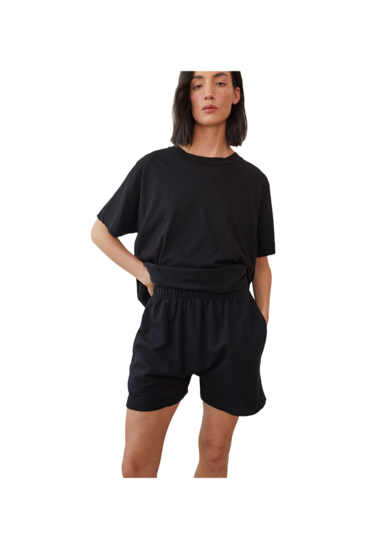 A person with shoulder-length dark hair is wearing a loose, black T-shirt tucked into Wol Hide's Cotton Easy Shorts in Black, which feature a high-rise elastic waistband. They stand with one hand on their hip against a plain, neutral background.