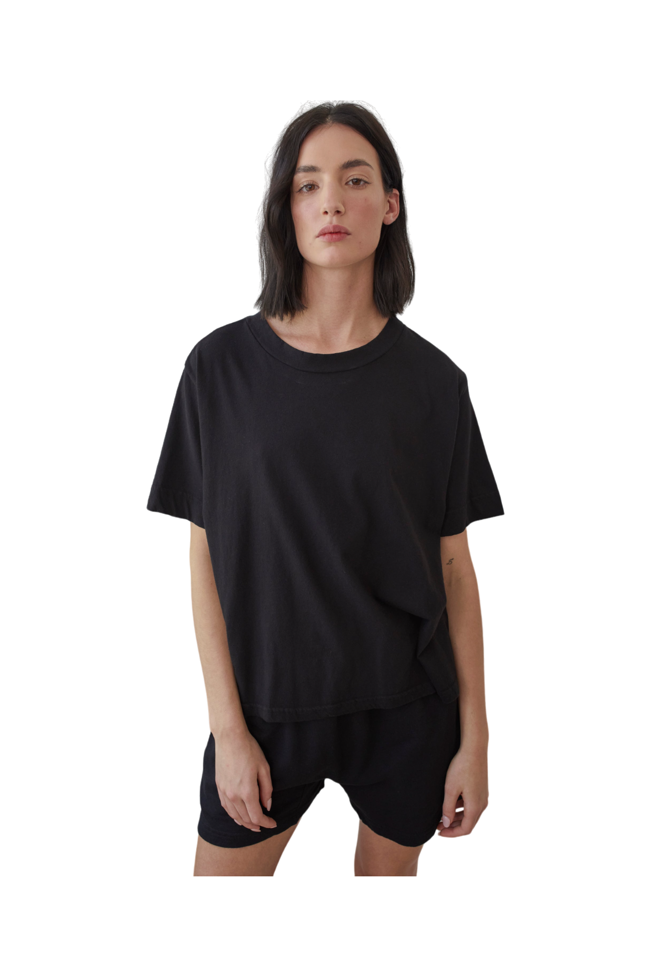 A person with shoulder-length dark hair stands against a plain white background, wearing a Wol Hide Organic Cotton Jersey Box Tee in Black and black shorts. Crafted from organic cotton and made in the USA, their outfit exudes both comfort and style.