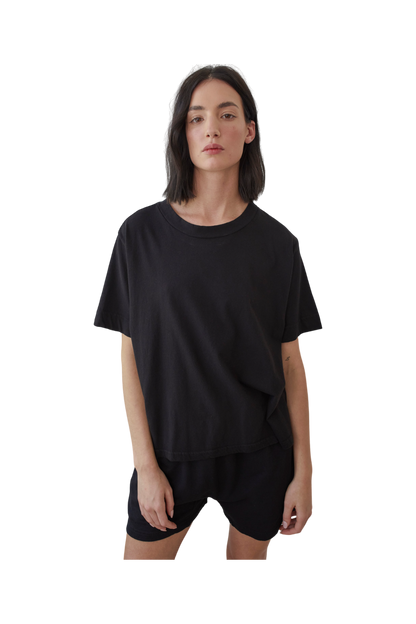 A person with shoulder-length dark hair stands against a plain white background, wearing a Wol Hide Organic Cotton Jersey Box Tee in Black and black shorts. Crafted from organic cotton and made in the USA, their outfit exudes both comfort and style.