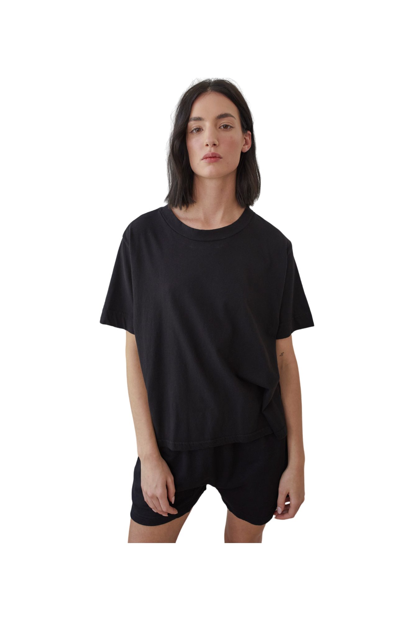 A person with shoulder-length dark hair wears Wol Hide's Organic Cotton Jersey Box Tee in black and shorts, made from organic cotton. Set against a plain white background, they look directly at the camera with a neutral expression. Made in the USA.