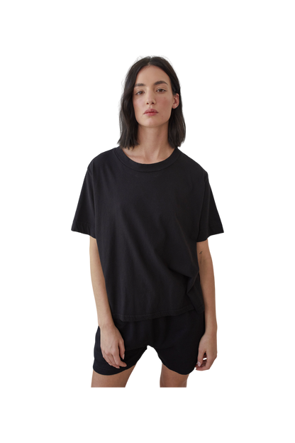 A person with shoulder-length dark hair wears Wol Hide's Organic Cotton Jersey Box Tee in black and shorts, made from organic cotton. Set against a plain white background, they look directly at the camera with a neutral expression. Made in the USA.