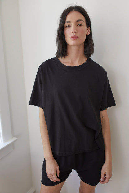 A person with shoulder-length dark hair is wearing a loose Organic Cotton Jersey Box Tee in Black by Wol Hide, made in the USA, and black shorts. They stand by a window in a well-lit room, sporting a neutral expression.