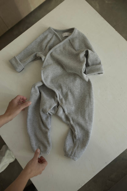 Hands are seen adjusting the APRÈS WRAP organic waffle, a light gray, long-sleeved newborn outfit from cabane childrenswear, on a white surface. This ethically handmade onesie showcases button snaps along the legs and a wrap-style front.