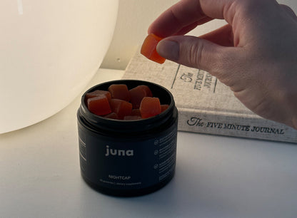 A hand reaches to take an orange gummy from a black container labeled "Nightcap Sleep Gummies" by Juna, a sleep aid filled with cubed gummies. In the background, there is a book titled "The Five Minute Journal," partially visible on a white surface. These functional botanicals support restorative sleep.