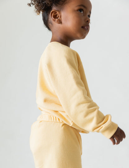 A child with curly hair stands in profile wearing the Lil' Classic Crewneck from all the babies, a long-sleeve, light yellow outfit crafted from organic French terry cloth. The child's head is turned slightly to the right against a plain, light background. This cozy crewneck is sure to become a beloved closet staple.