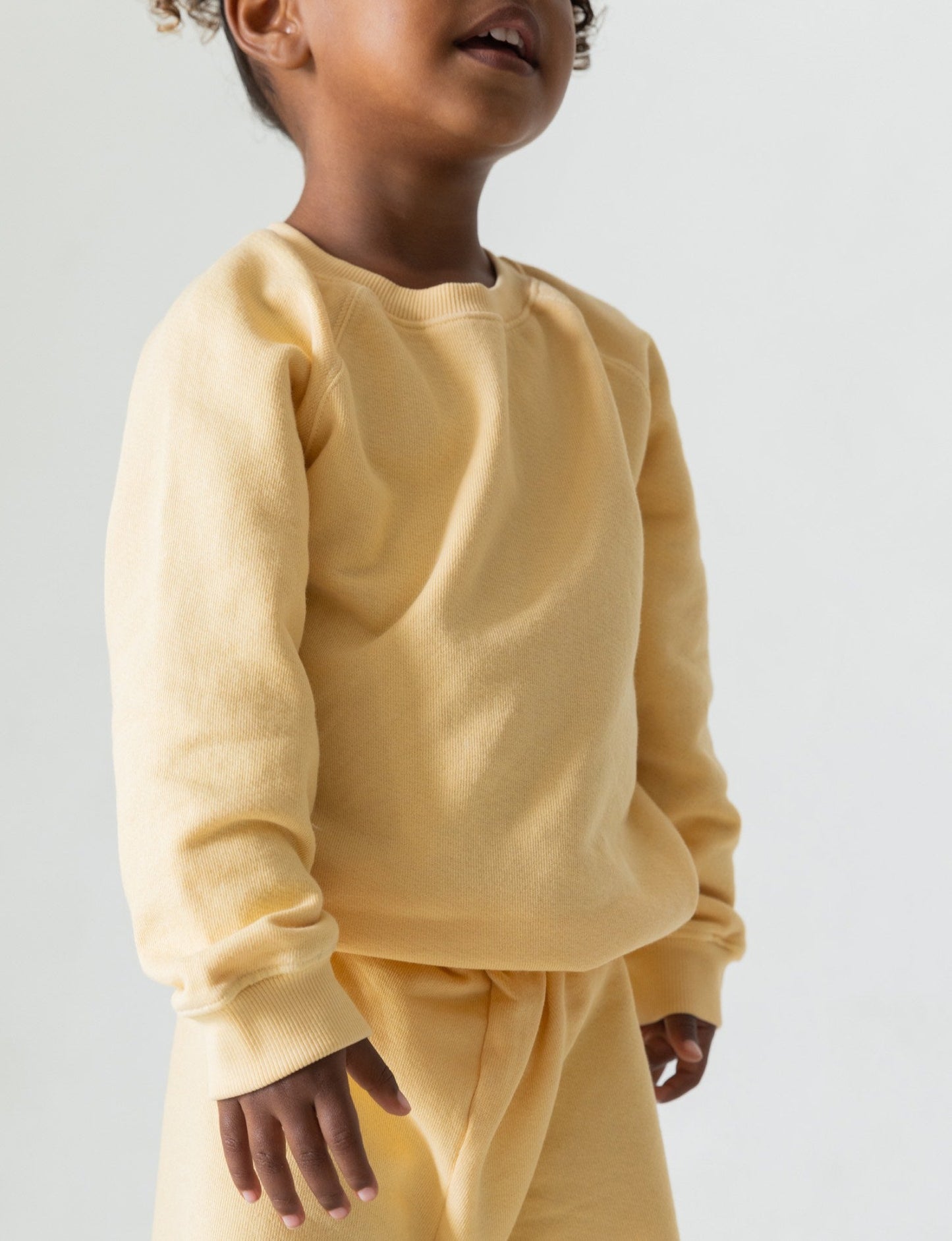 A young child is pictured from the shoulders down, wearing The Lil' Classic Crewneck and matching pants set by all the babies, made from organic French terry cloth in a light yellow hue. The image is set against a plain, light background. The child's head is slightly tilted up, with visible lips suggesting a happy expression.