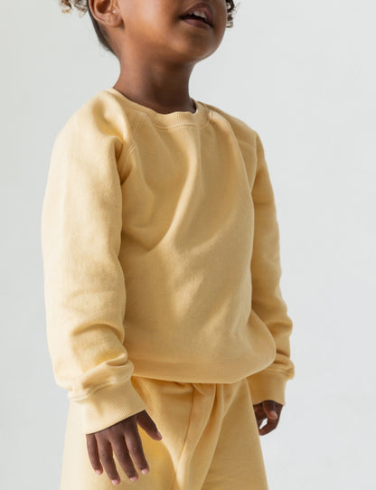 A young child is pictured from the shoulders down, wearing The Lil' Classic Crewneck and matching pants set by all the babies, made from organic French terry cloth in a light yellow hue. The image is set against a plain, light background. The child's head is slightly tilted up, with visible lips suggesting a happy expression.