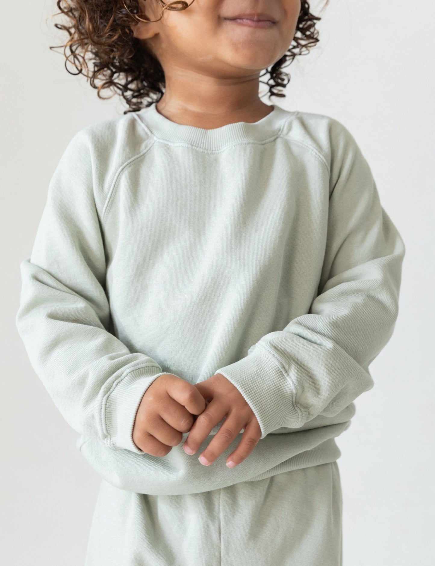 A young child with curly hair is smiling and wearing "The Lil' Classic Crewneck" in light grey by all the babies, paired with matching pants. They are standing against a plain white background with their hands clasped in front of them, showcasing the cozy closet staple made from organic French terry cloth.