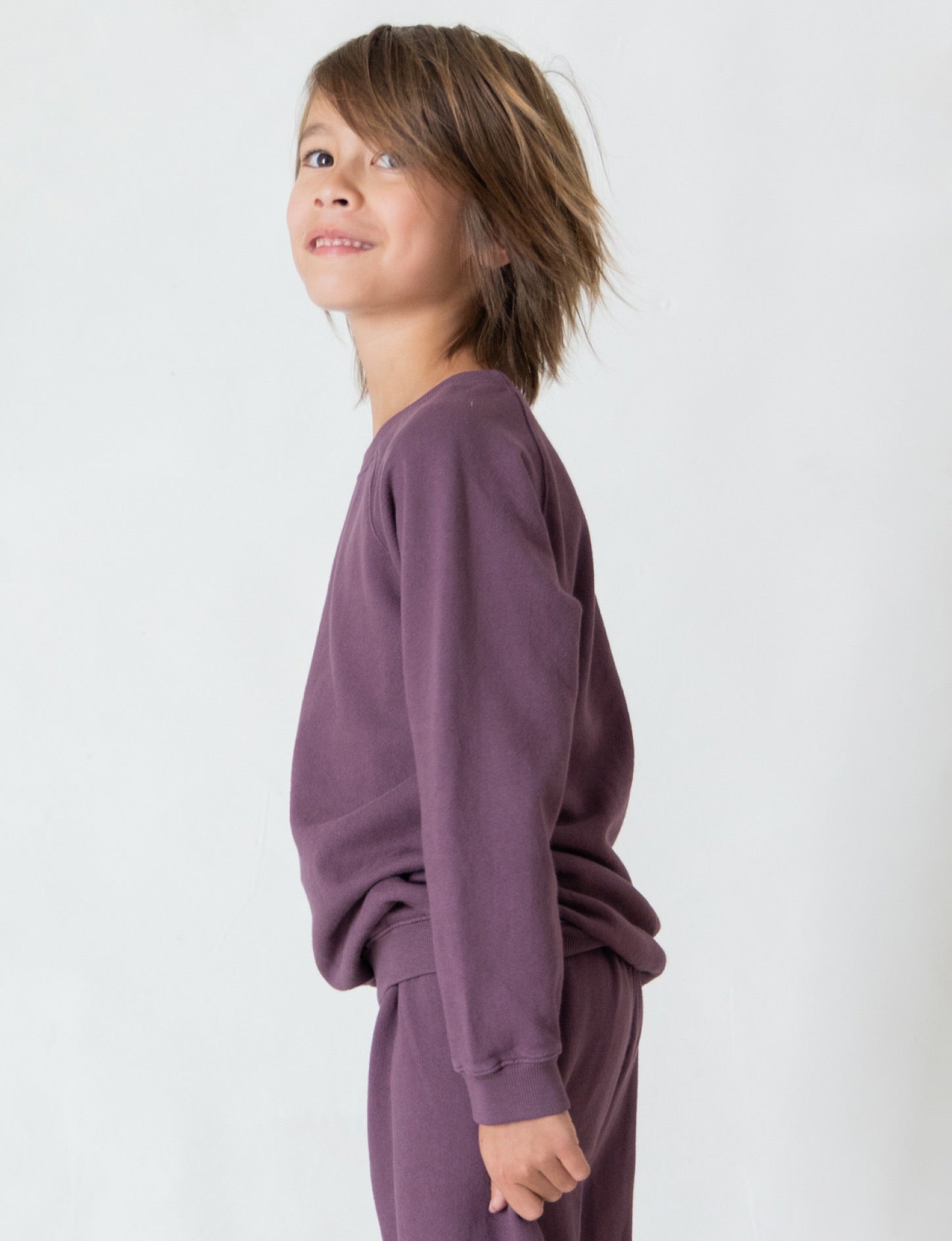 A young child with shoulder-length brown hair is smiling and looking slightly upwards. They are wearing The Lil' Classic Crewneck, a matching purple long-sleeve shirt and pants set made from organic French terry cloth by all the babies. The background is plain and light-colored, showcasing the cozy closet staple outfit perfectly.