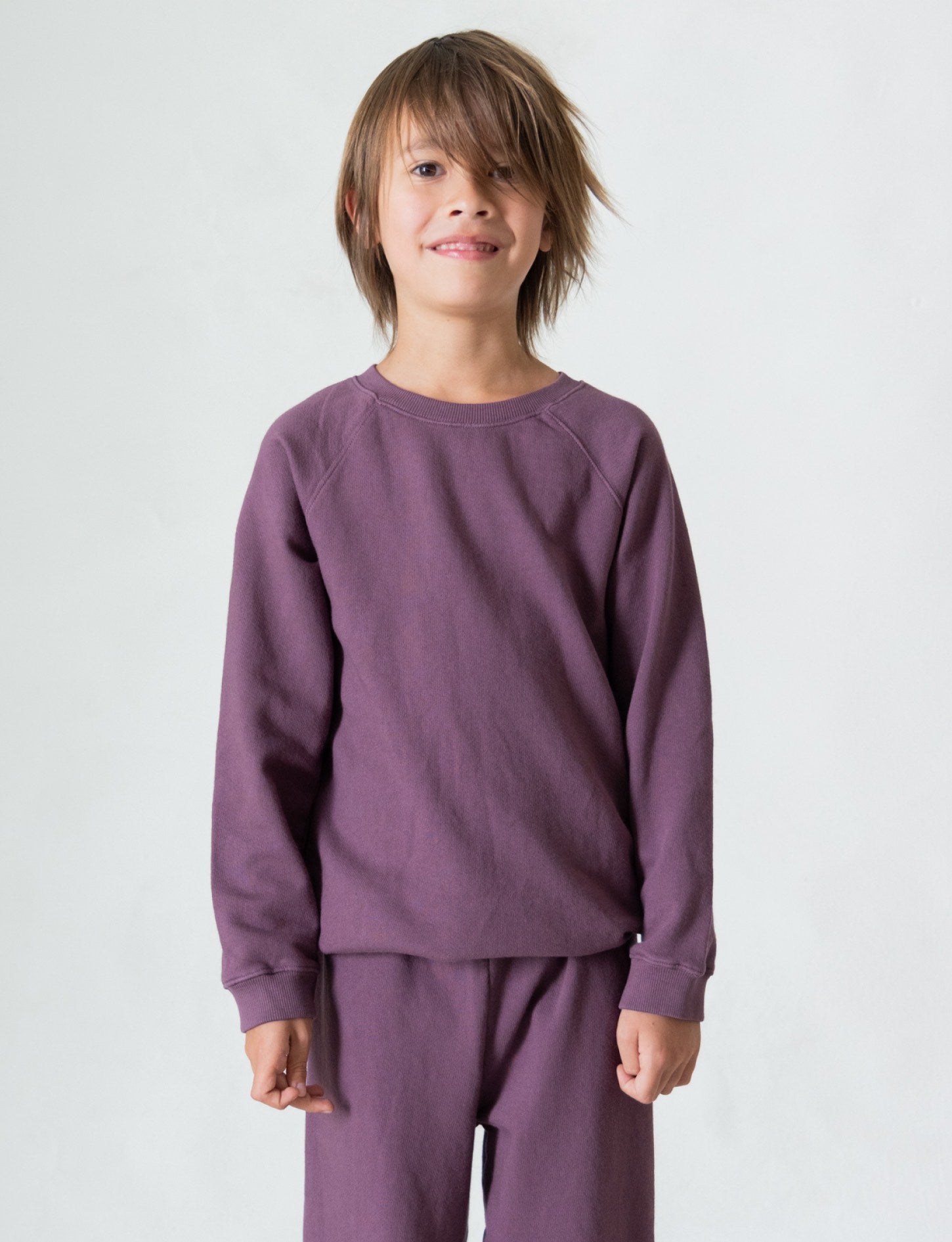 A young child with shaggy brown hair is standing and smiling at the camera, dressed in The Lil' Classic Crewneck by all the babies and matching pants made from organic French terry cloth. The plain white background enhances the timeless appeal of this cozy closet staple.