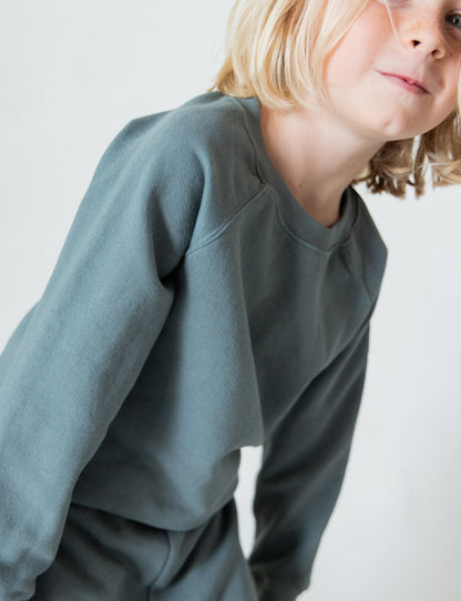 A child with long, blonde hair wearing The Lil' Classic Crewneck and matching pants from the brand all the babies, made of organic French terry cloth. The child appears to be in motion, leaning slightly forward and smiling towards the camera against a plain, light-colored background.