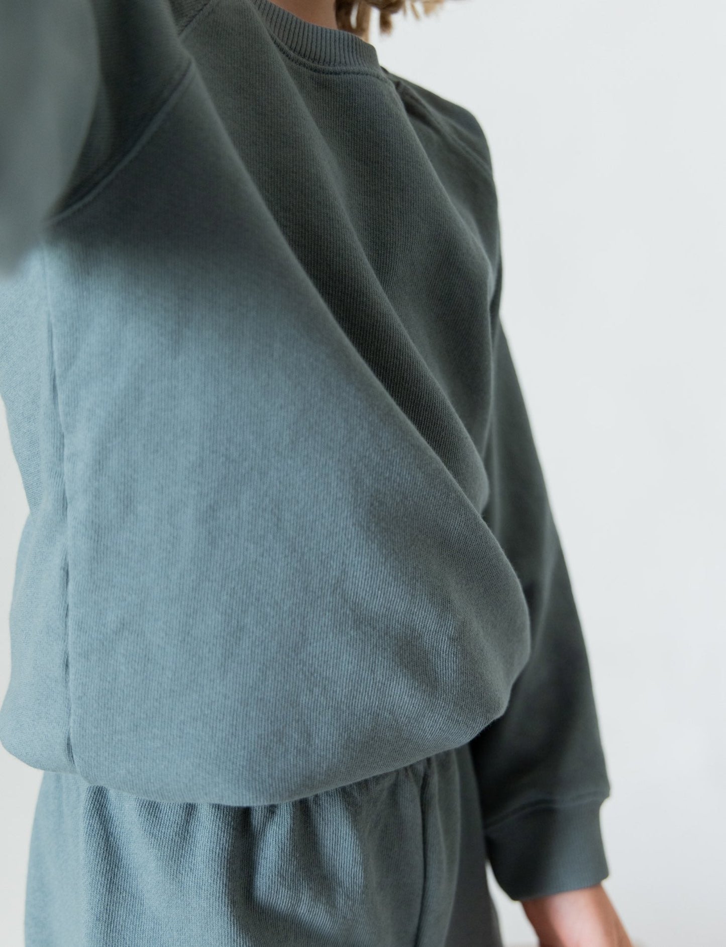 A person wearing the Lil' Classic Crewneck and matching pants from all the babies, made of organic French terry cloth, is partially visible, with the upper torso and parts of the arms and shoulders in focus. This cozy closet staple stands out against a plain white background.