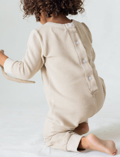 A young child with curly hair is kneeling on the floor, facing away from the camera. They are wearing "The Thermal Onesie" by all the babies, which is a cream-colored, textured organic onesie with buttons down the back. The child is holding a soft toy in their right hand.