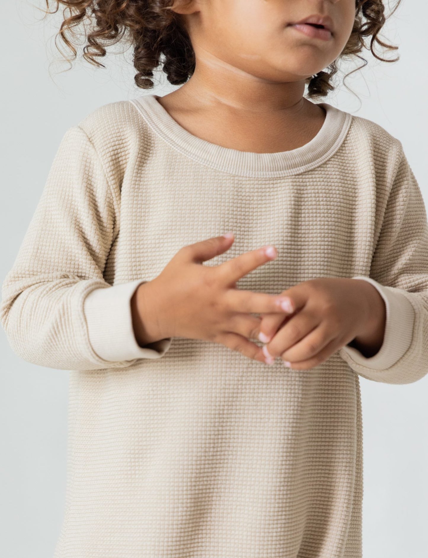 A young child with curly hair stands against a plain background, wearing The Thermal Onesie in light beige from all the babies. They have their hands held loosely together in front of them, while their face is partially out of the frame.