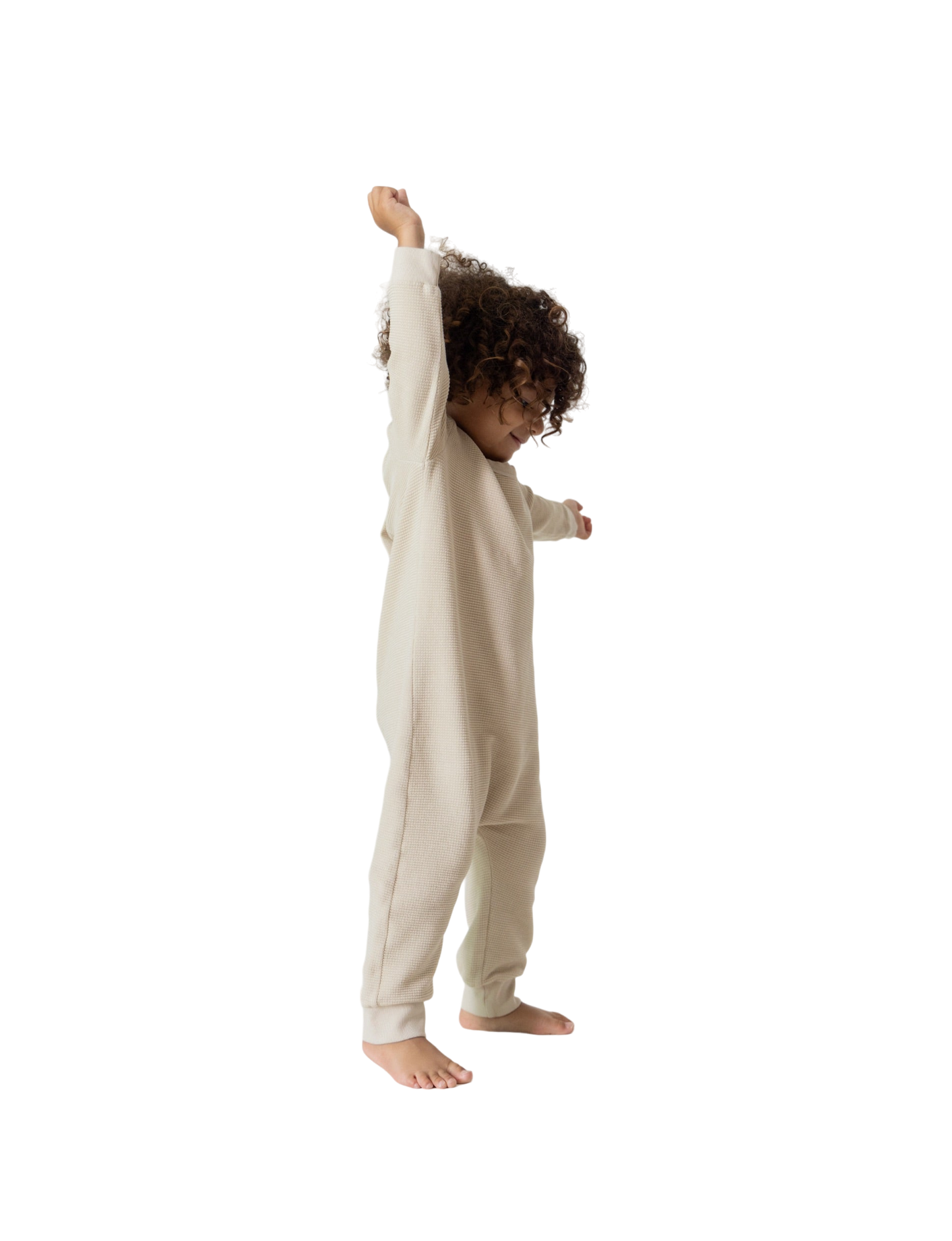 A young child with curly hair stands barefoot, joyfully stretching their left arm upwards, clad in all the babies' full-length, long-sleeved beige Thermal Onesie. The background is plain white.