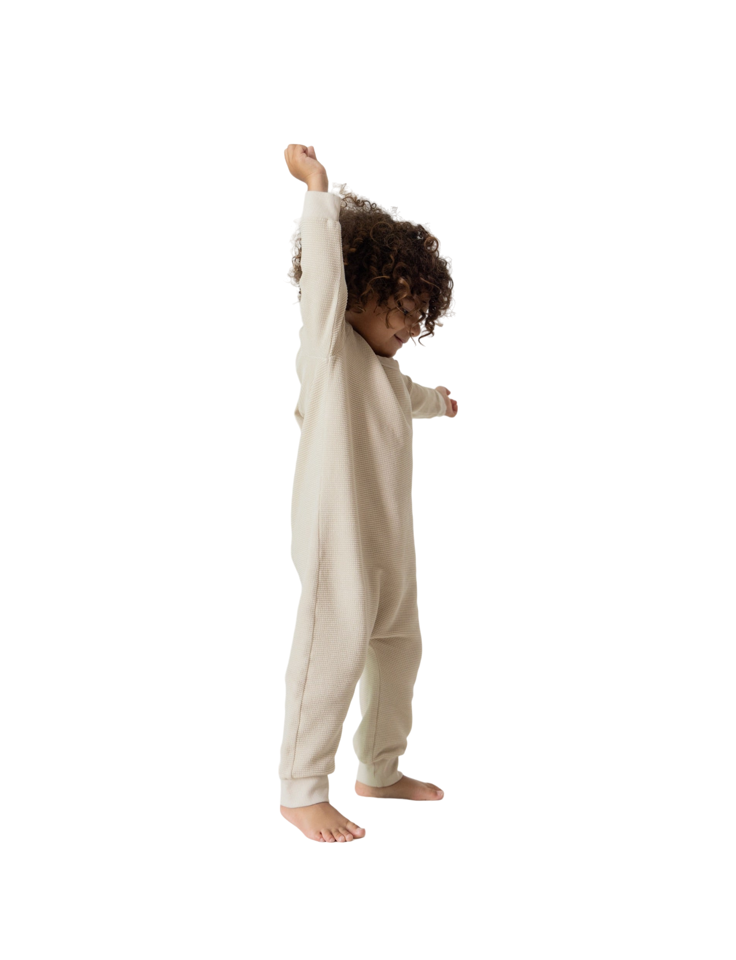 A young child with curly hair stands barefoot, joyfully stretching their left arm upwards, clad in all the babies' full-length, long-sleeved beige Thermal Onesie. The background is plain white.