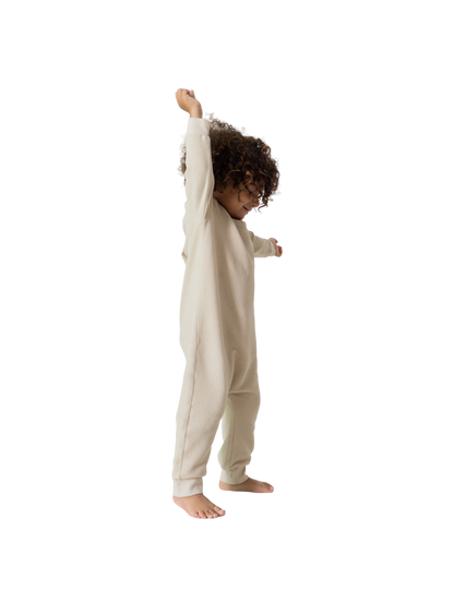 A young child with curly hair stands barefoot, joyfully stretching their left arm upwards, clad in all the babies' full-length, long-sleeved beige Thermal Onesie. The background is plain white.