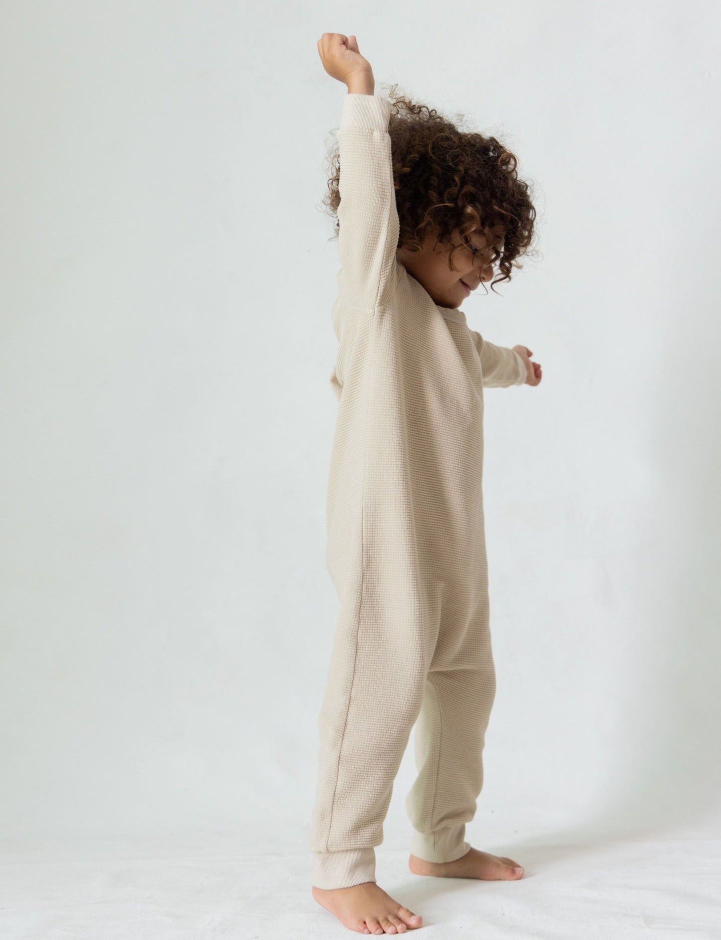 A small child with curly hair is standing barefoot on a white floor against a white background. The child is wearing The Thermal Onesie from all the babies, a beige long-sleeve organic onesie, and has their arms raised as if they are dancing or stretching.