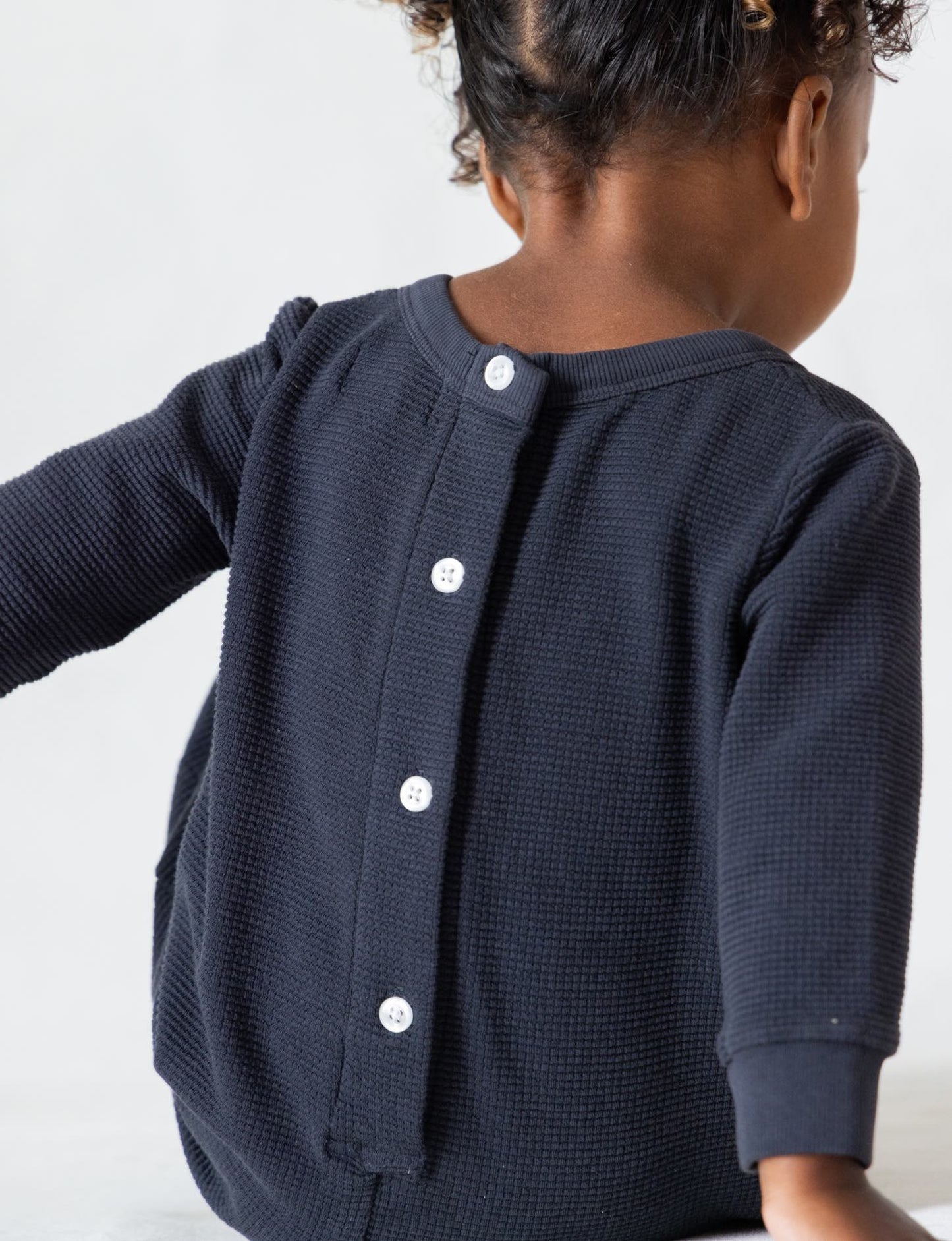 A young child with curly hair is pictured from the back, wearing "The Thermal Onesie" by all the babies – a long-sleeved, dark blue knit top that resembles a cozy thermal onesie with a row of five white buttons down the back. The child is seated and facing away from the camera against a plain, light-colored background.