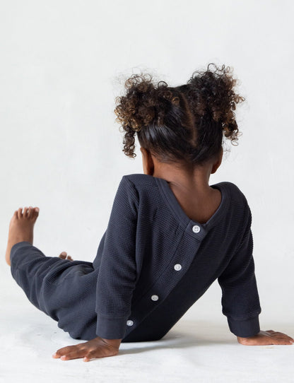 A small child with curly hair sits on the floor with their back turned to the camera. The child is wearing The Thermal Onesie by all the babies, a dark, long-sleeved outfit featuring buttons down the back. The background is plain and light-colored.